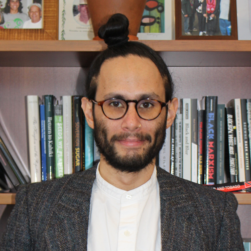 Uahikea Maile against a bookshelf wearing glasses, a white collarless shirt and a dark blazer